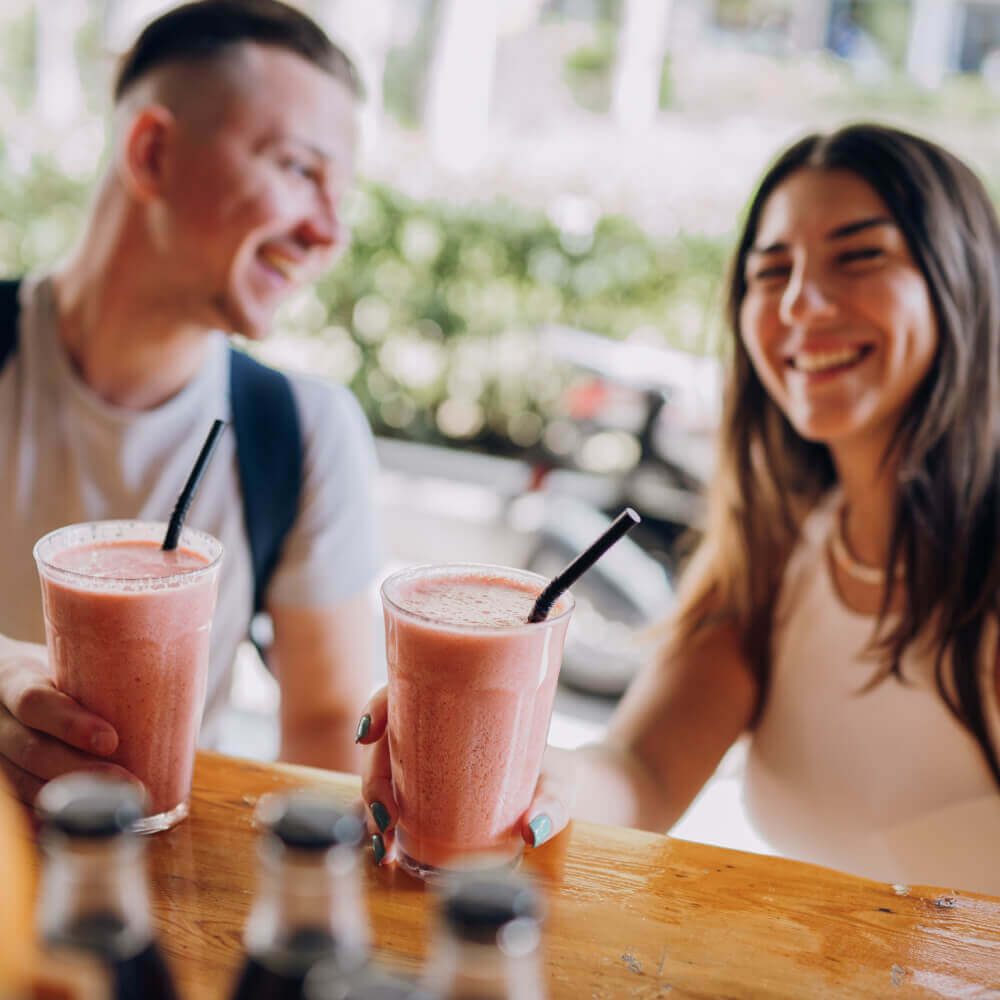 Two people enjoy pink smoothies at Village Clubs Camelback, seated by a sunny window with lush greenery and bottles on the table.