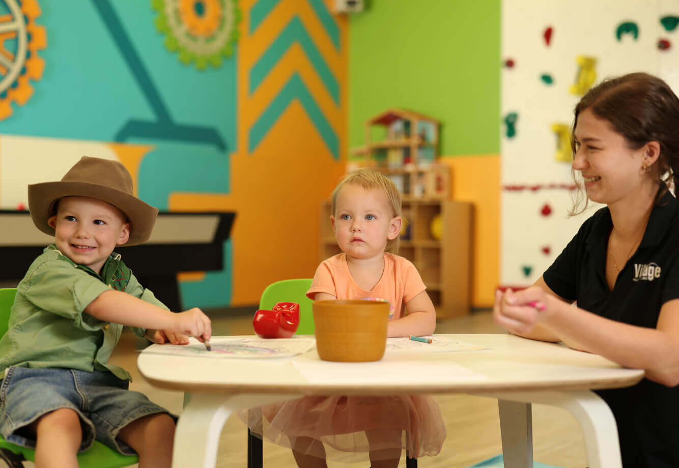 At Village Clubs DC Ranch, two kids color at a small table as a smiling woman supervises in a bright, colorful playroom.