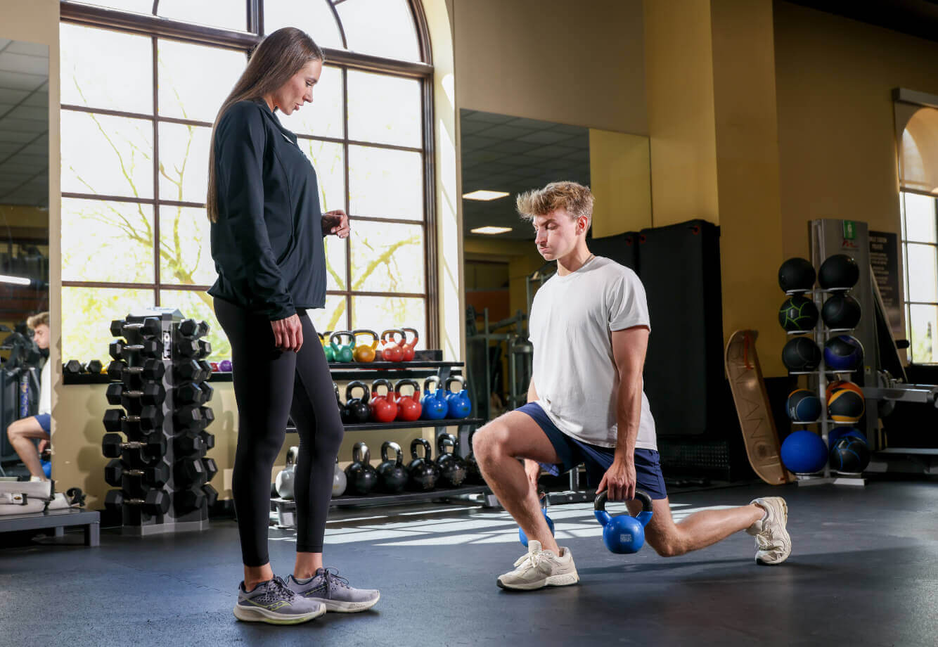 At Village Clubs Camelback, a man lunges with a kettlebell as a trainer supervises in the gym with equipment and large windows.
