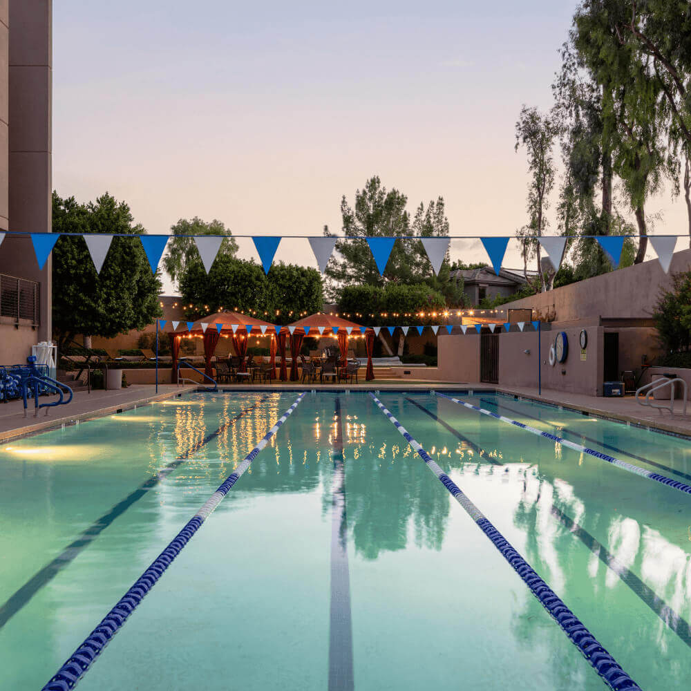 Village Clubs Camelback outdoor pool at sunset with lane dividers, pennant flags, and string-lit seating surrounded by trees.