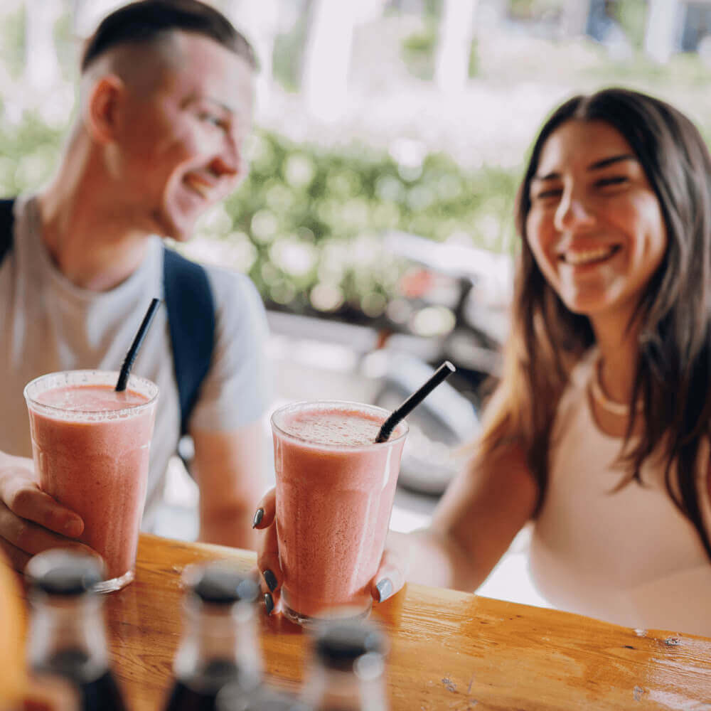 Two people enjoy pink smoothies at Village Clubs Camelback, seated outdoors at a wooden table with bottles in the foreground.