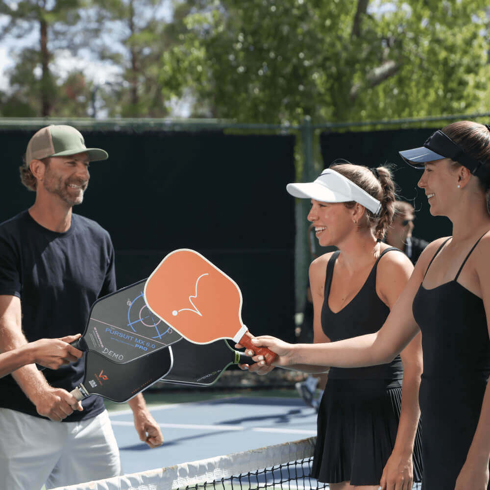 Members at Village Clubs Camelback play pickleball outdoors, smiling at the net with paddles; court fence and trees behind them.
