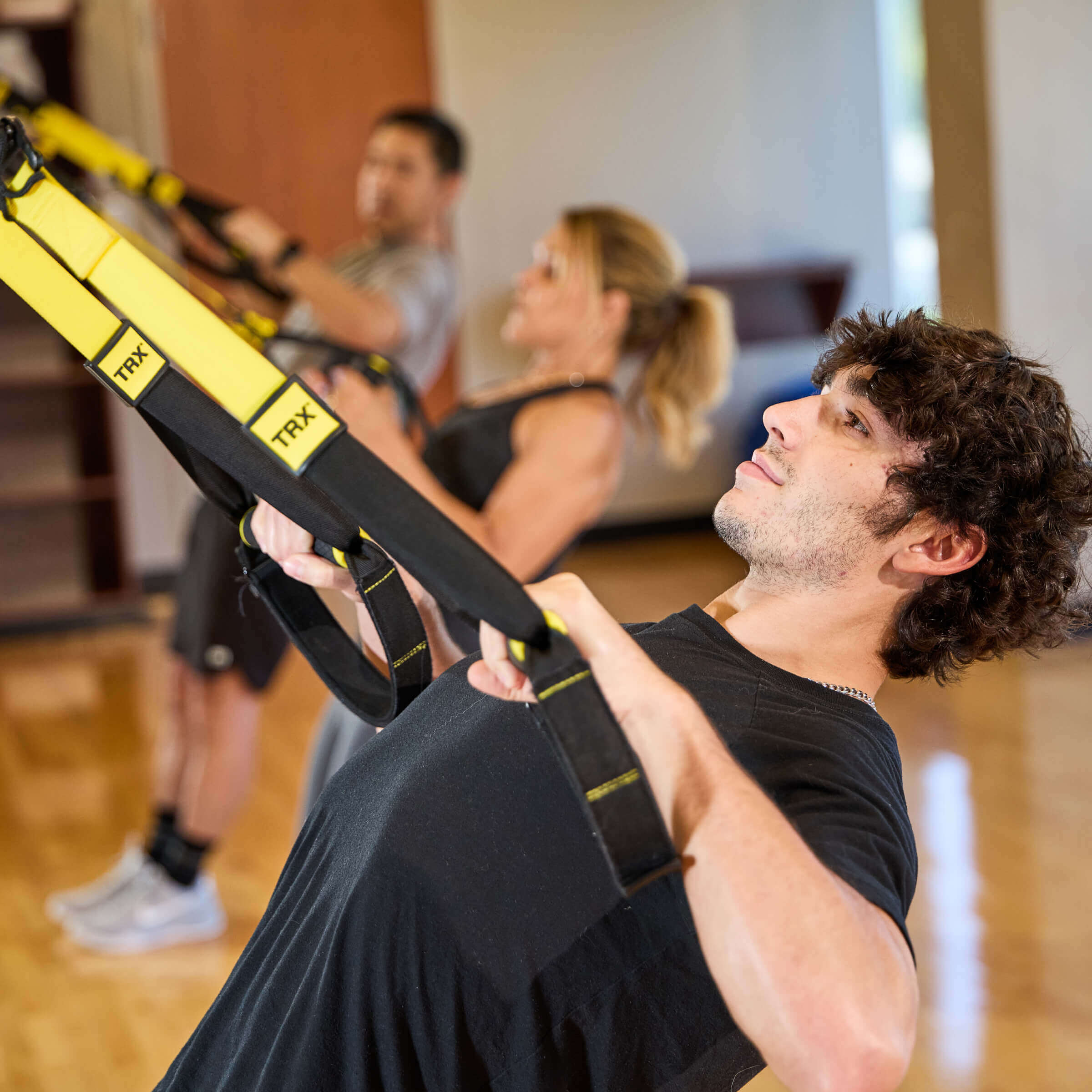 Members use yellow and black TRX suspension trainers at Village Clubs Camelback, in a bright room with wood floors.