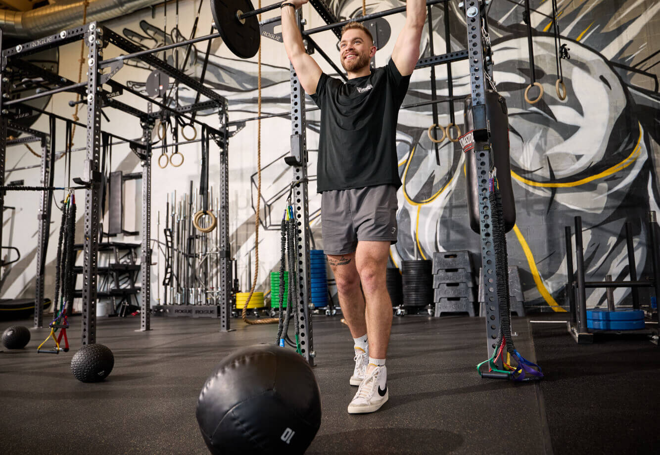 A man lifts a barbell overhead at Village Clubs Camelback gym, surrounded by equipment and a colorful graffiti mural on the wall.
