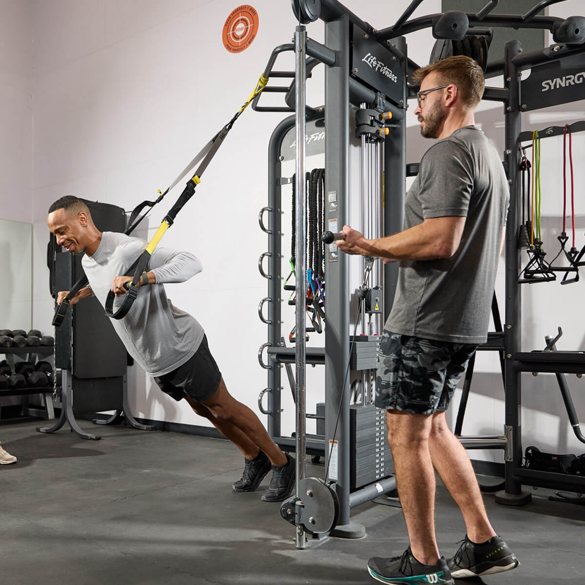 Two men training at Village Clubs Camelback gym—one on suspension straps, another on a cable machine; equipment fills the background.