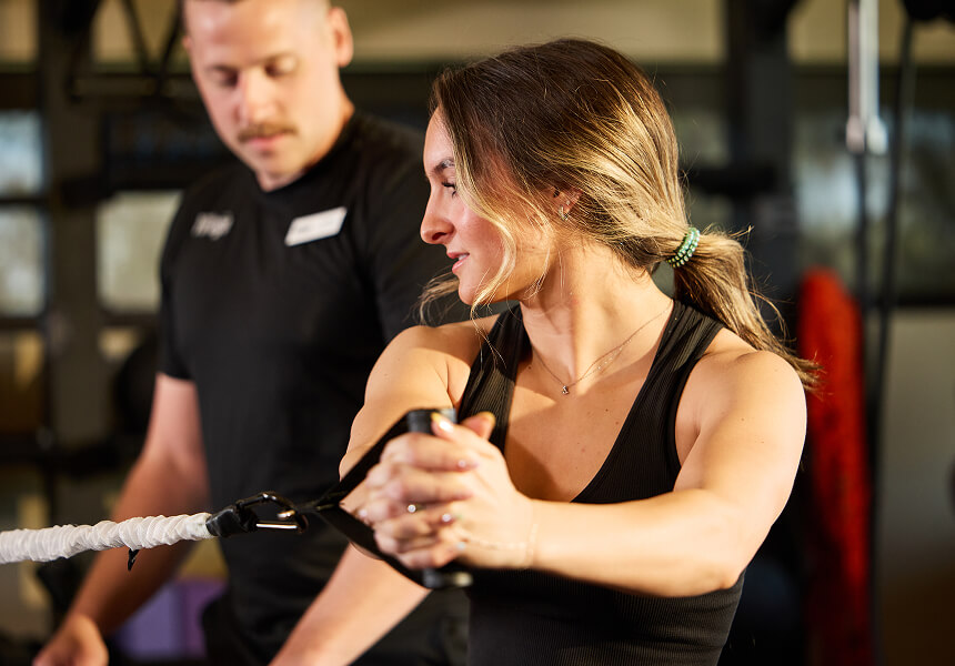 A woman works out with a resistance band at Village Clubs Camelback, guided by a fitness trainer in the gym.