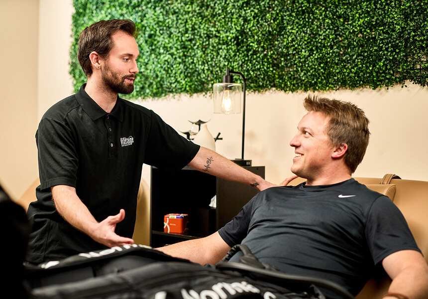 Village Clubs Camelback staff member assists a guest using leg compression therapy sleeves in a relaxing lounge with greenery.