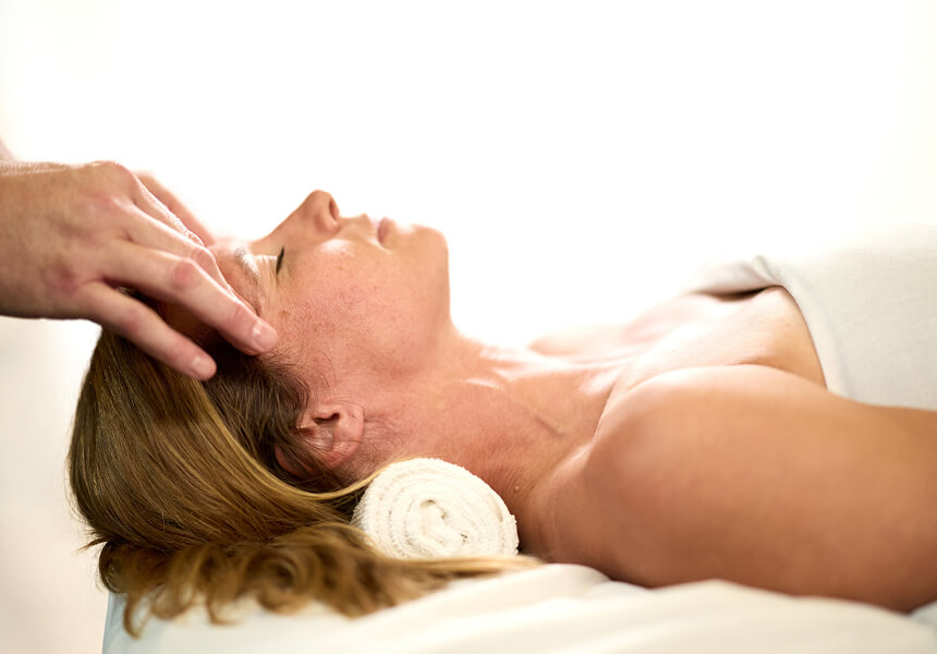 A woman enjoys a relaxing head massage at Village Clubs Camelback, lying with a towel under her neck in a bright, serene space.