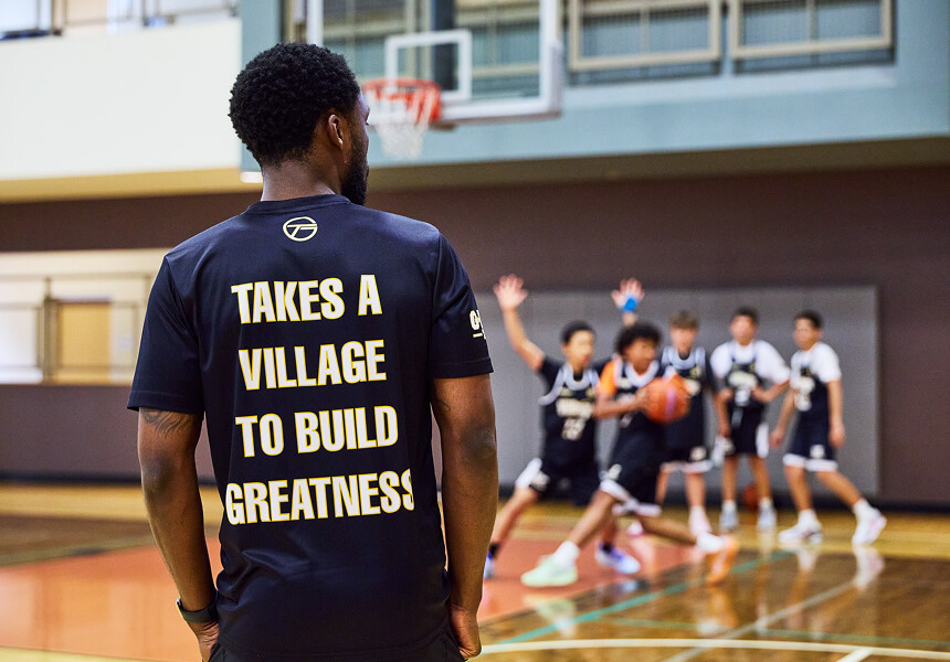 At Village Clubs Camelback, a coach wearing a TAKES A VILLAGE TO BUILD GREATNESS shirt guides youth basketball practice on court.