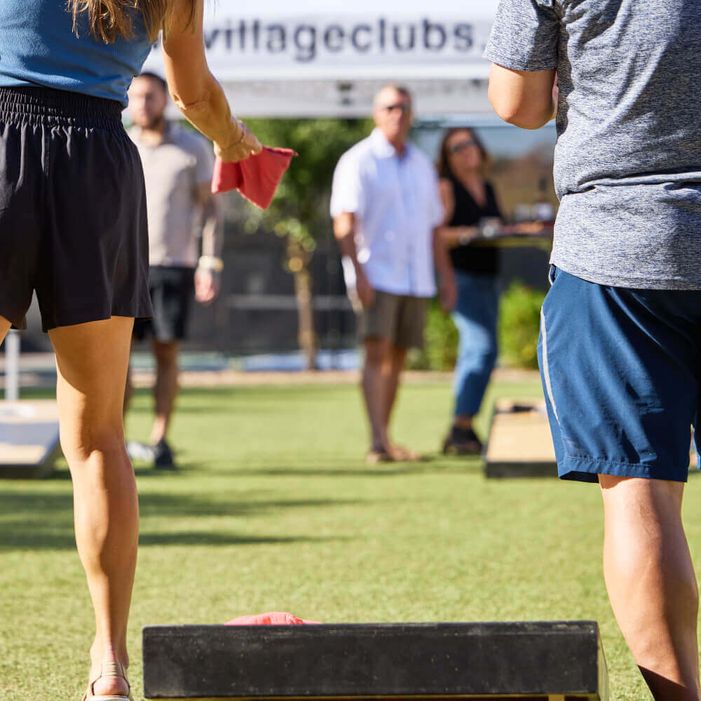 Members enjoy a sunny game of cornhole on the grass at Village Clubs Camelback in Arizona, with others socializing nearby.