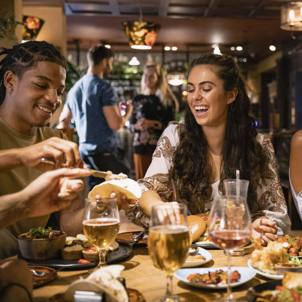 Friends enjoying food and drinks at Village Clubs Camelback, with a lively restaurant atmosphere and others socializing nearby.