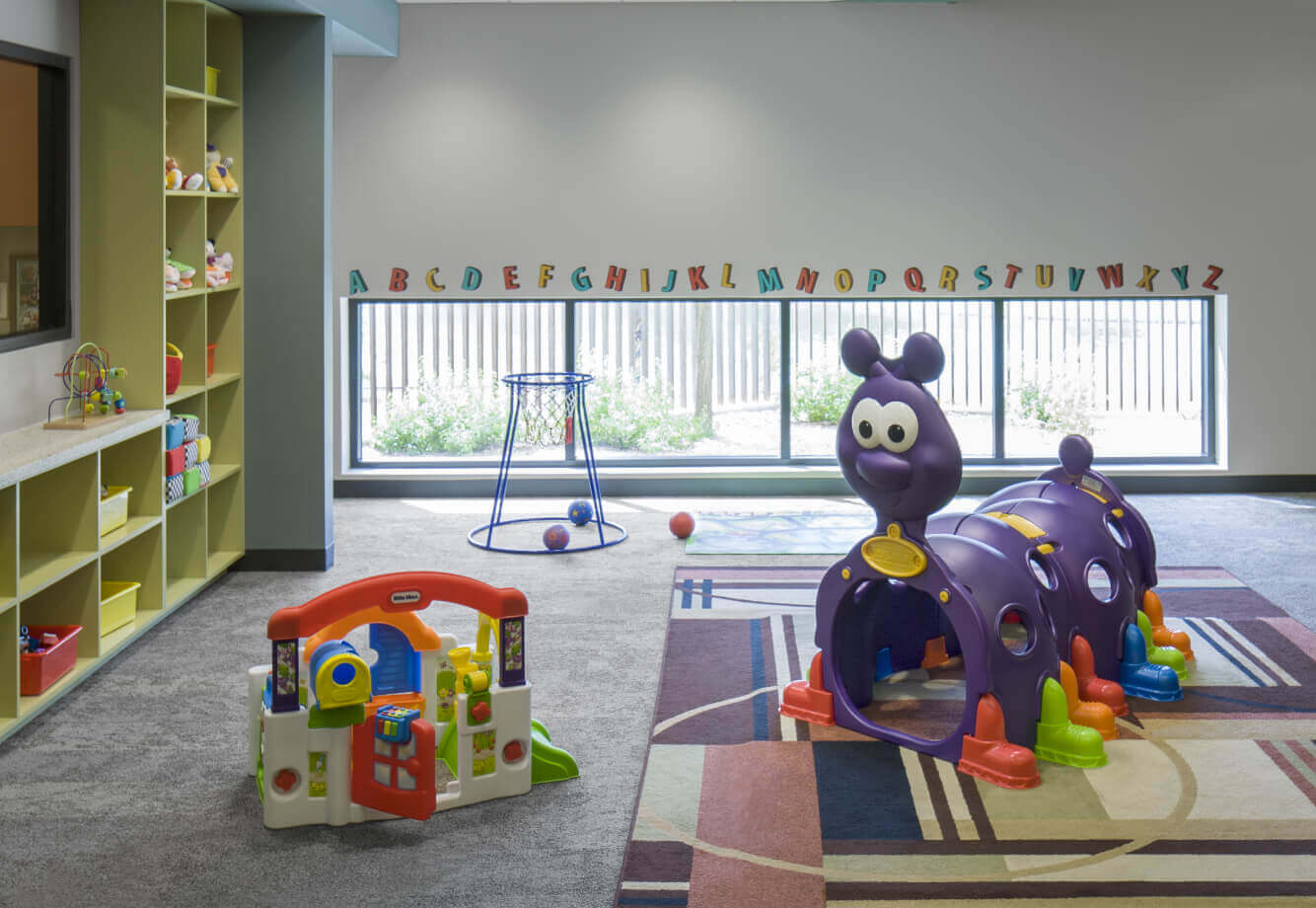Village Clubs Camelback playroom with toys, activity center, caterpillar tunnel, and sunlight through large windows.