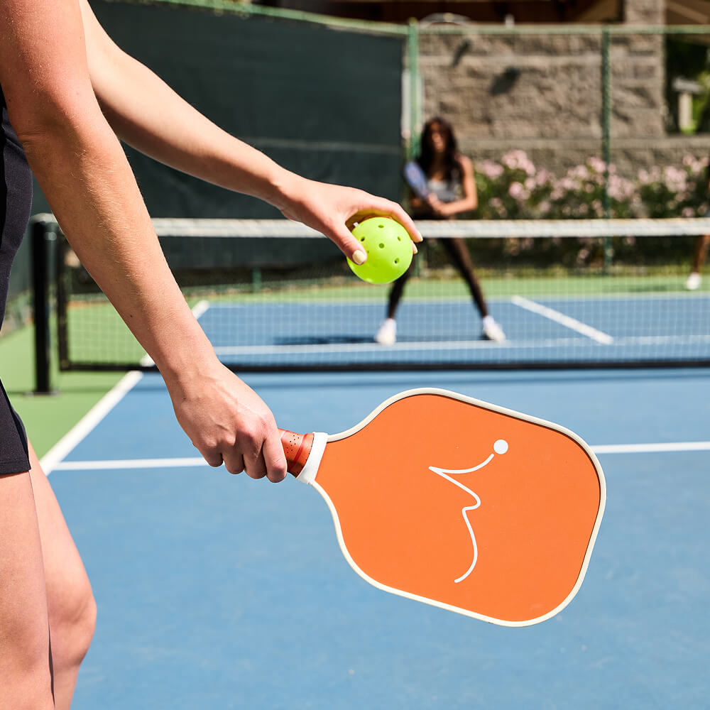 At Village Clubs Camelback in Arizona, a player prepares to serve pickleball on an outdoor court while their opponent stands ready.