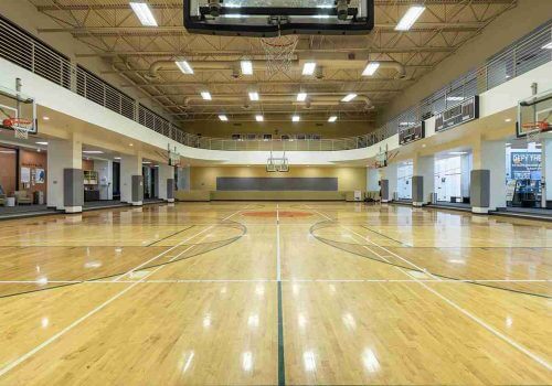 Village Clubs Camelback indoor basketball court with polished wood floors, high ceilings, balcony seating, and dual hoops.