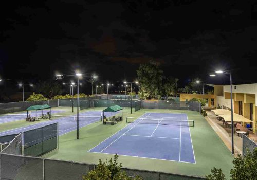 Village Clubs Camelback outdoor tennis courts at night with green benches, covered seating, and adjacent clubhouse for spectators.