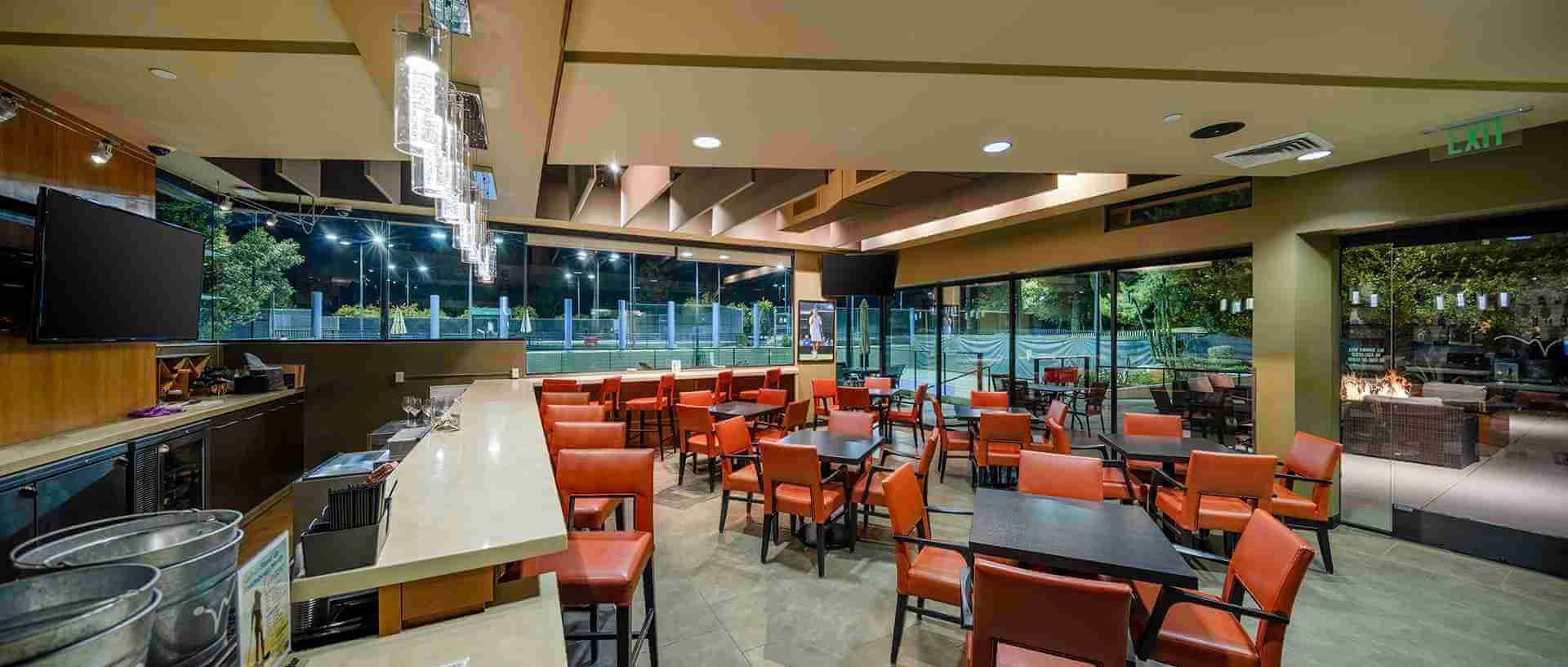 Village Clubs Camelback restaurant with red chairs, bar, tennis court views, and outdoor fire pit visible through large windows.