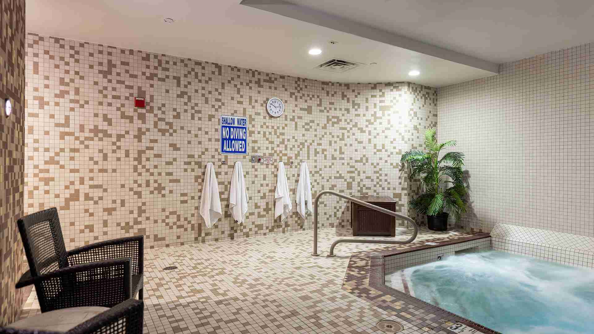 Village Clubs indoor hot tub at DC Ranch with bubbling water, mosaic tiles, towels, chairs, potted plant, clock, and safety sign.