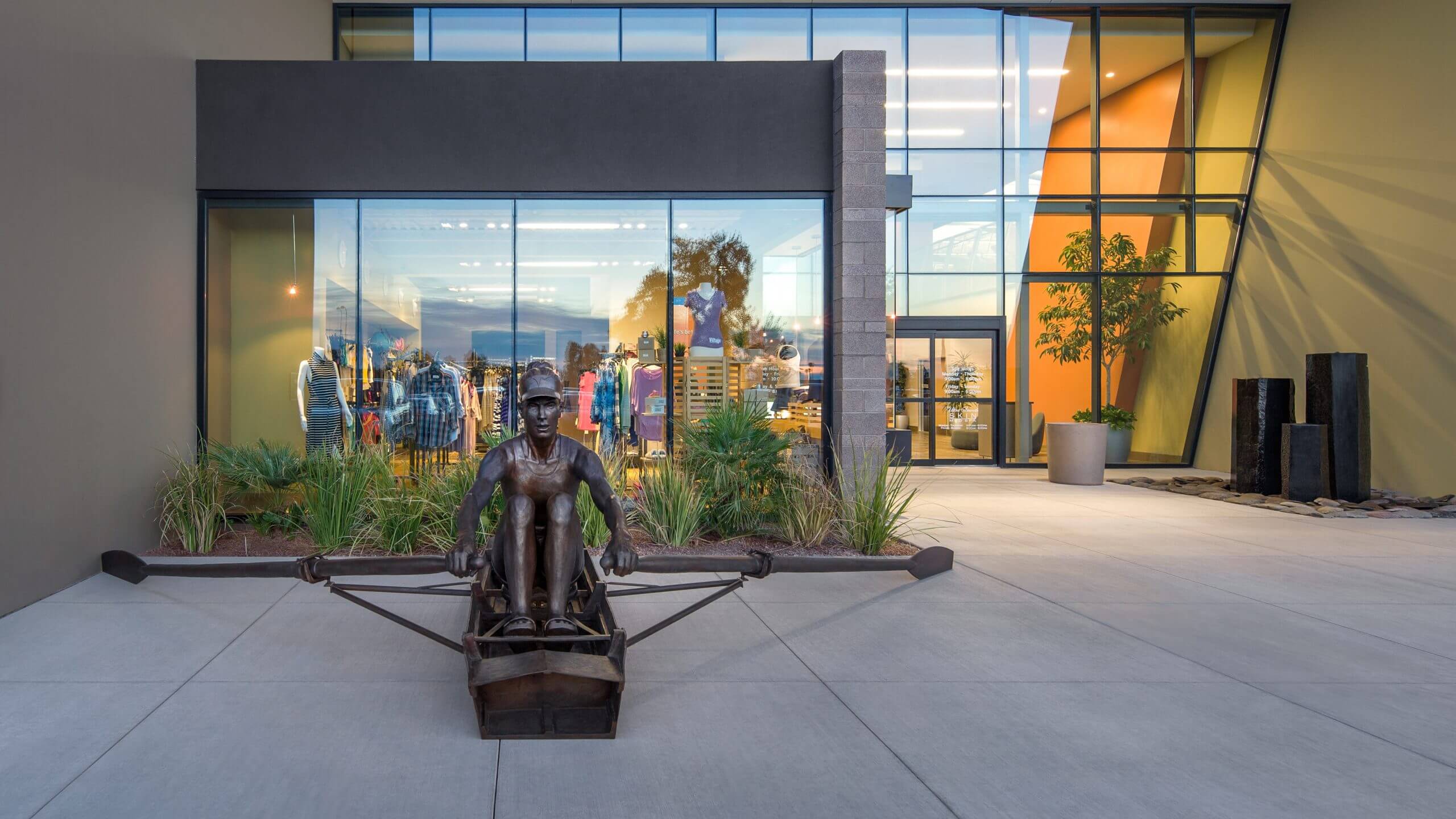 Bronze rowing sculpture at Village Clubs Ocotillo with glass windows showcasing athletic wear mannequins and a plant-lined entrance.