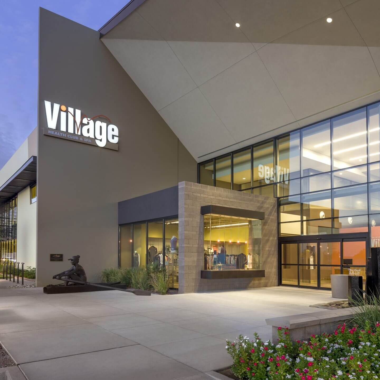 Village Clubs Ocotillo fitness center at dusk with glass windows, statue by entrance, and landscaped walkway.