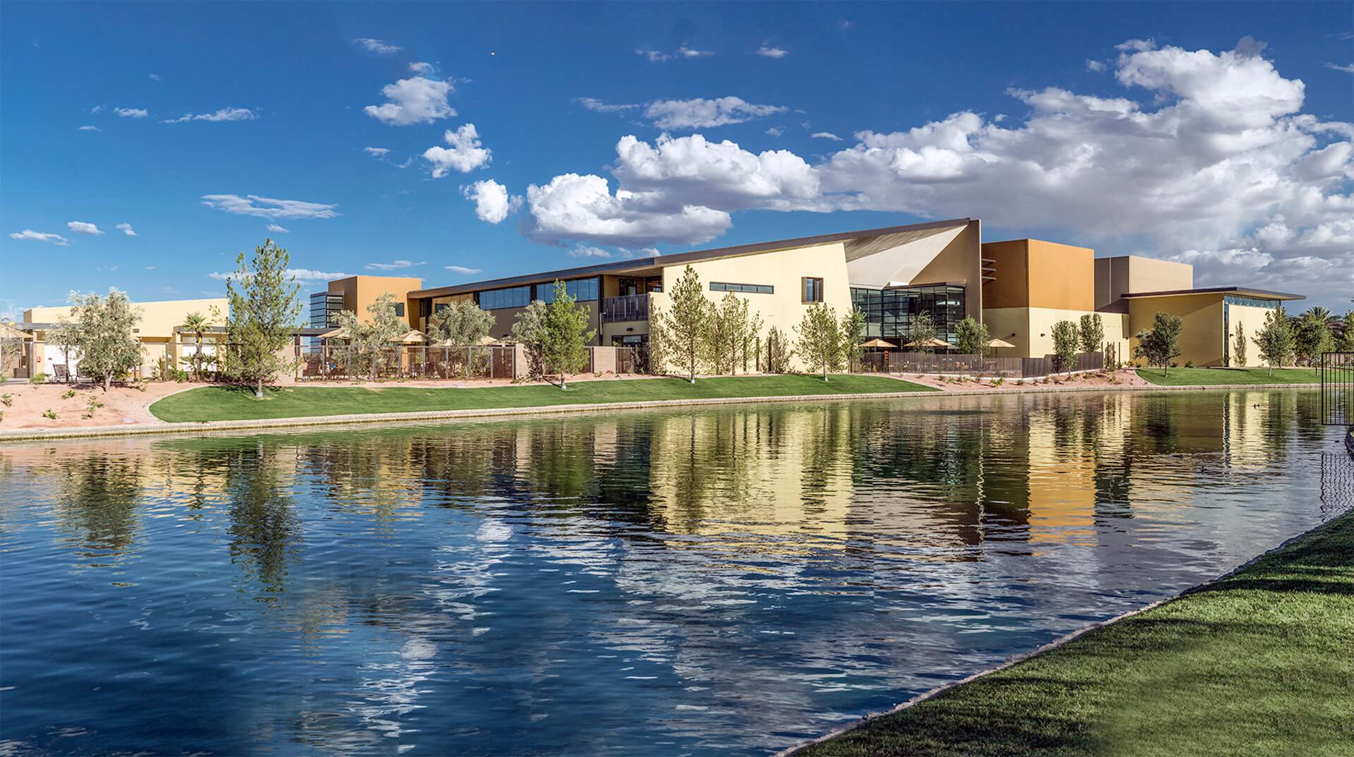 Village Clubs at Ocotillo features a modern, angular building by a lake, with lush landscaping and blue skies in Arizona.