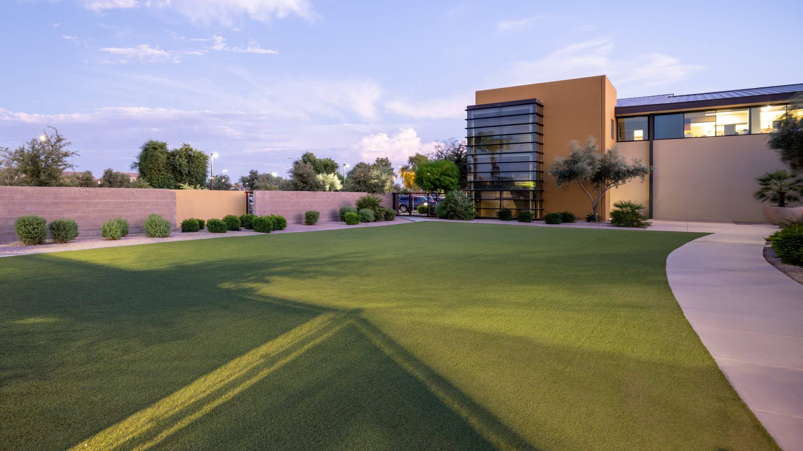 Village Clubs at Ocotillo features a modern glass building overlooking a manicured lawn with desert landscaping at dusk.