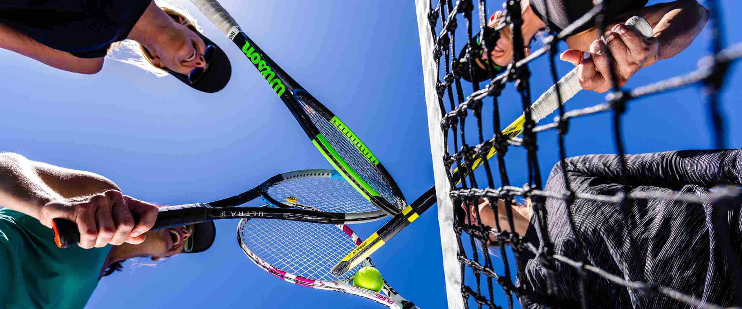 Players on a Village Clubs DC Ranch tennis court touch racquets over the net under a clear blue Arizona sky, tennis ball nearby.