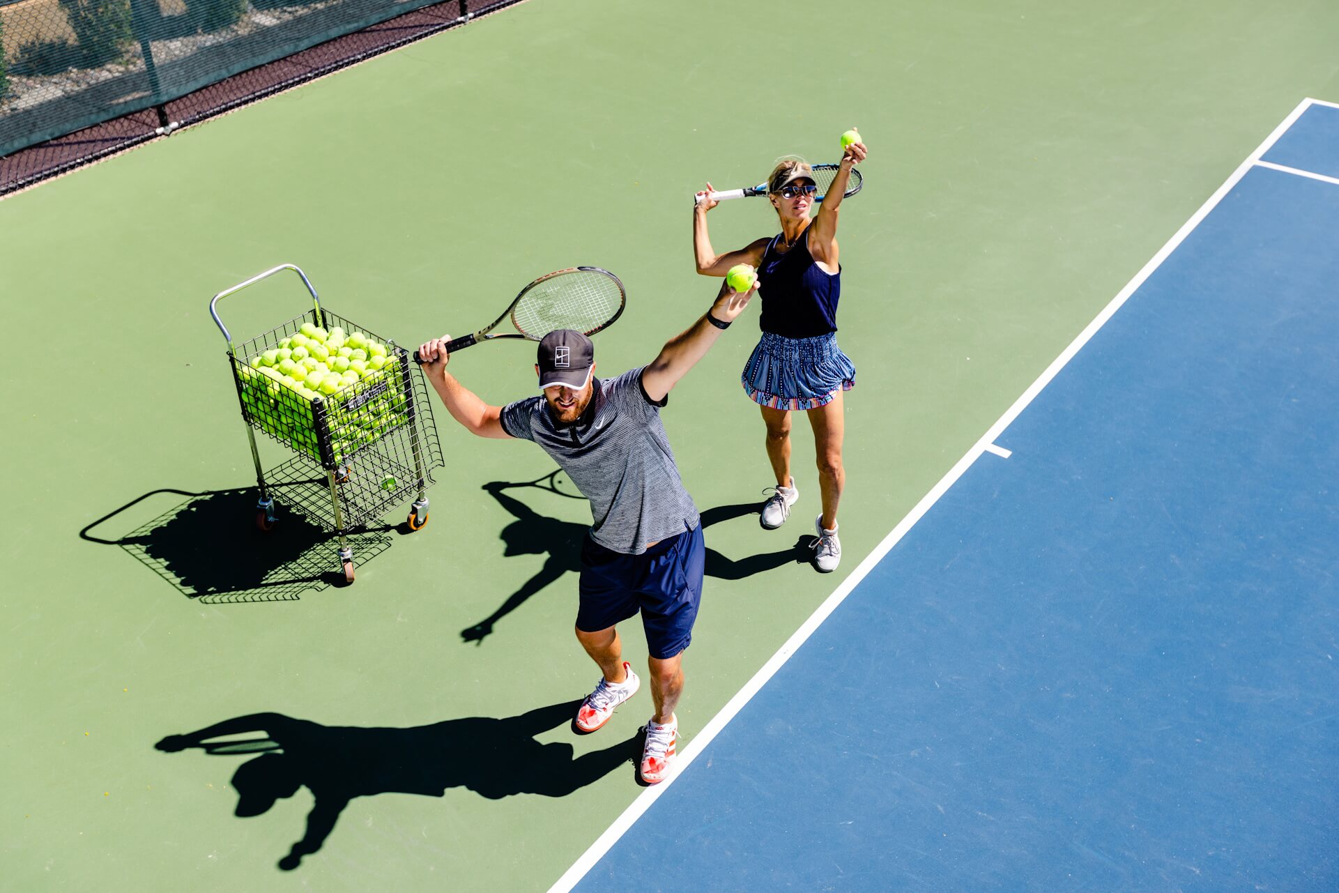 Two players serve on a Village Clubs tennis court at DC Ranch, with a ball cart nearby and clear court lines in an overhead view.