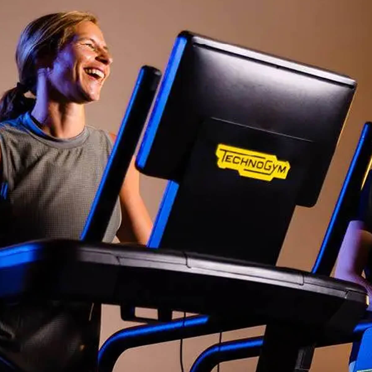A woman enjoys running on a Technogym treadmill at Village Clubs Camelback, smiling at a digital screen in a warmly lit gym.
