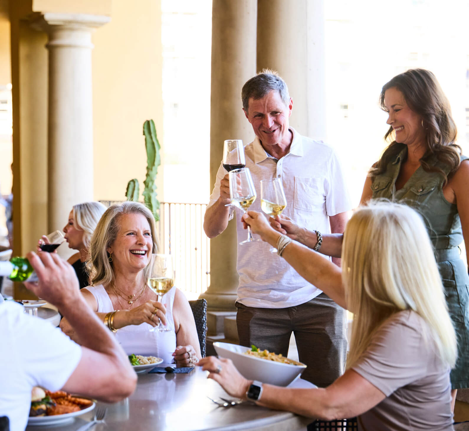 Adults gather outdoors at Village Clubs Camelback, smiling and toasting with wine amid pillars and cactus in a lively setting.