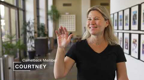 A smiling woman waves in a brightly lit Village Clubs Camelback hallway lined with framed photos. Text reads: Camelback Member Courtney.