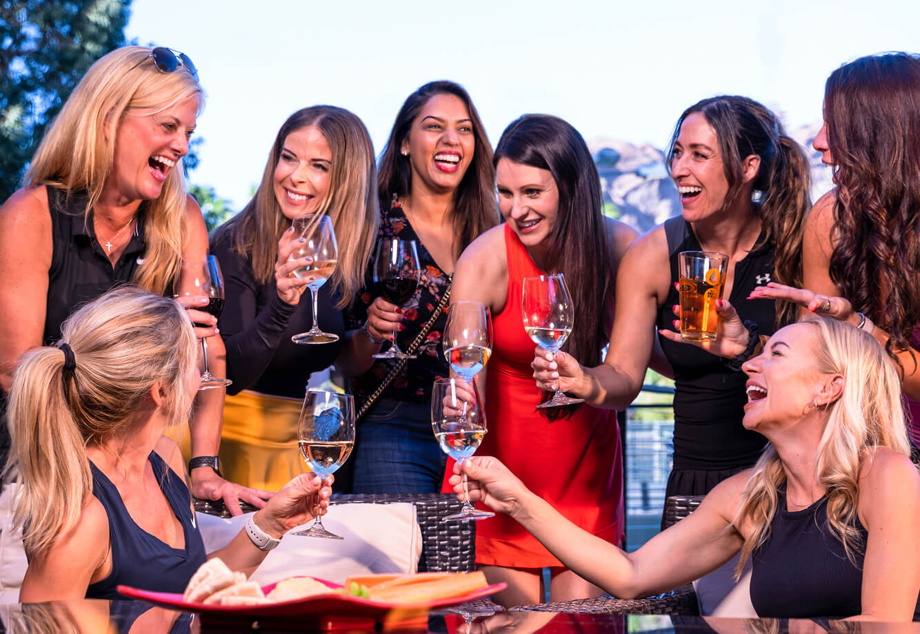 Women enjoy drinks and laughter at a Village Clubs Camelback outdoor gathering on a sunny day, celebrating community.
