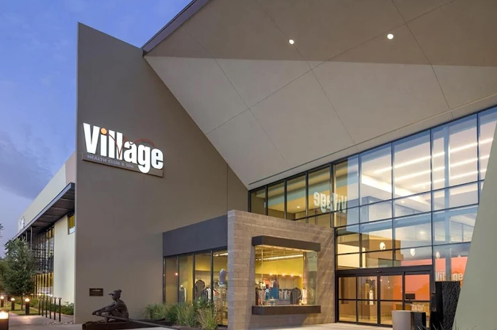 Village Clubs Camelback in Arizona with modern glass facade, metal sculpture, and plant-filled entrance under a clear sky.