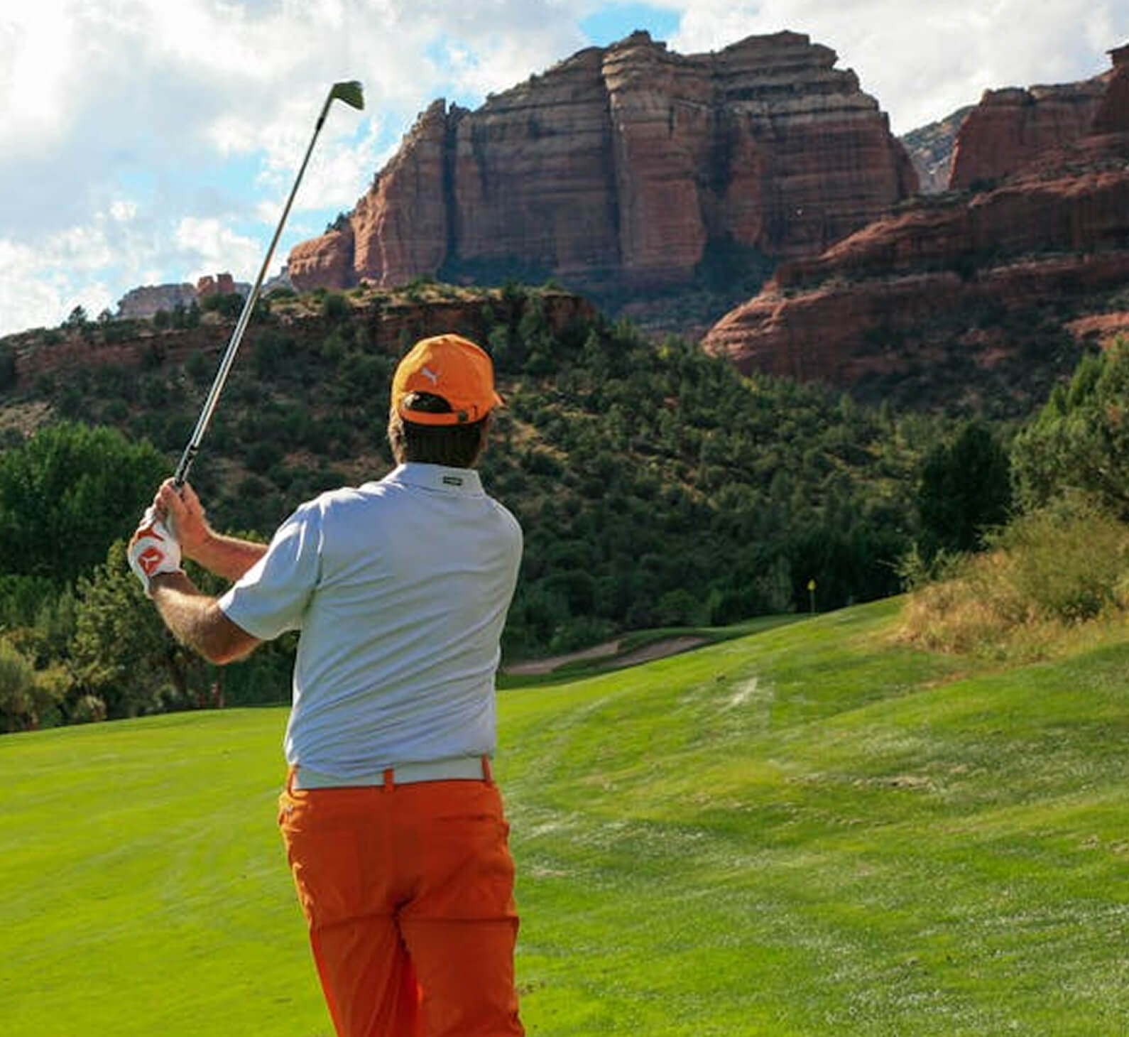 A golfer swings on a lush Village Clubs Camelback fairway, framed by red rocks and trees beneath a partly cloudy Arizona sky.