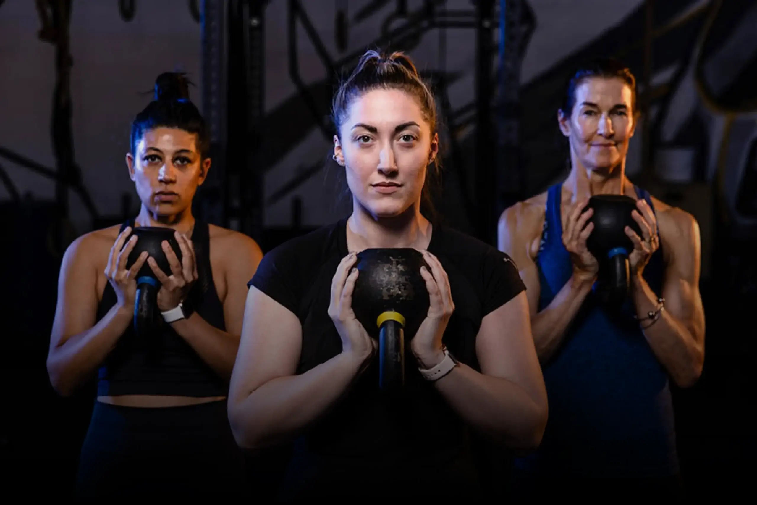 Three women at Village Clubs Camelback gym hold kettlebells in athletic wear, focusing on strength training in a modern facility.
