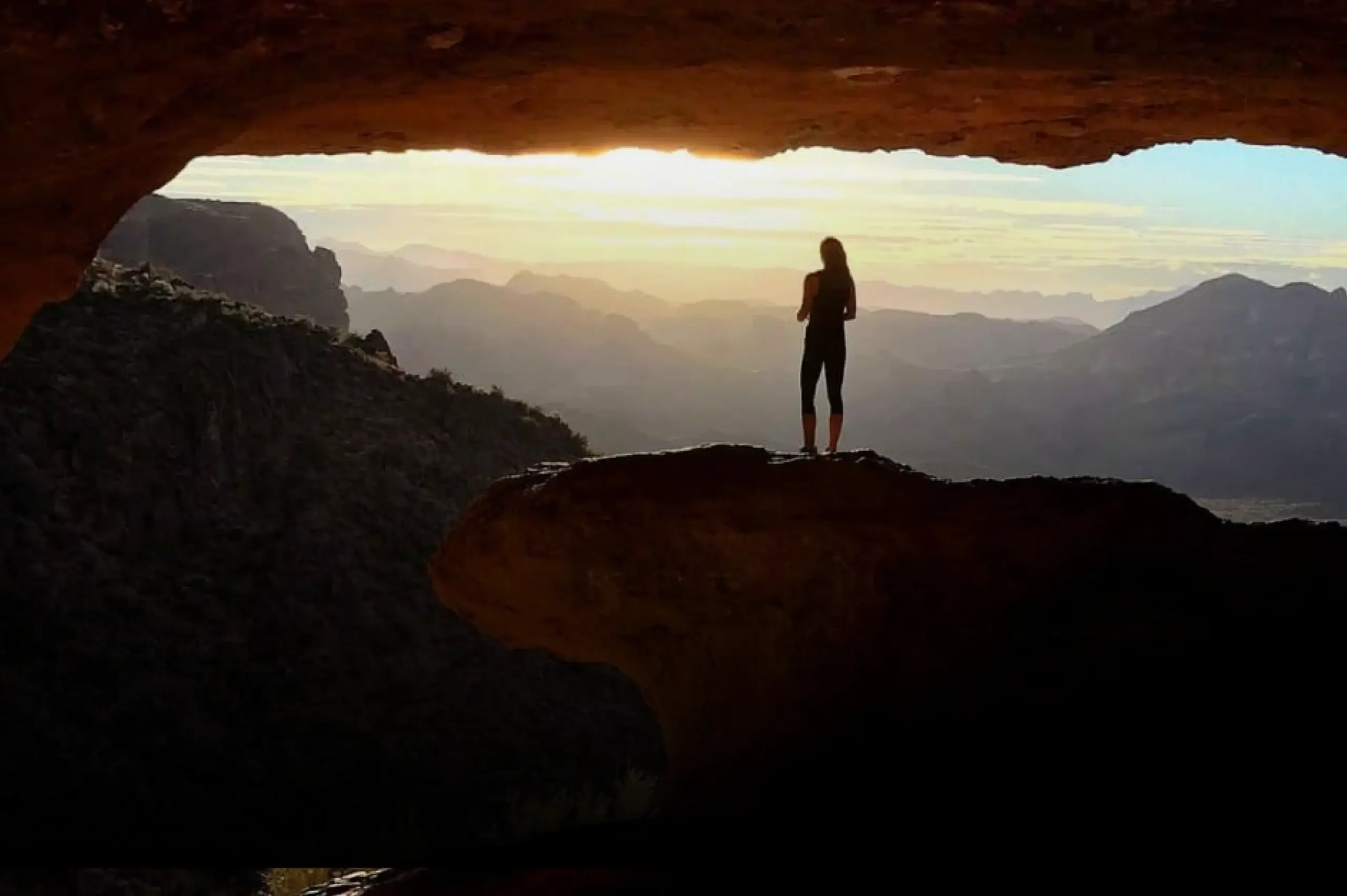 A person at Village Clubs Camelback stands on a rock in a cave, silhouetted by sunset over Arizona mountains and a glowing sky.