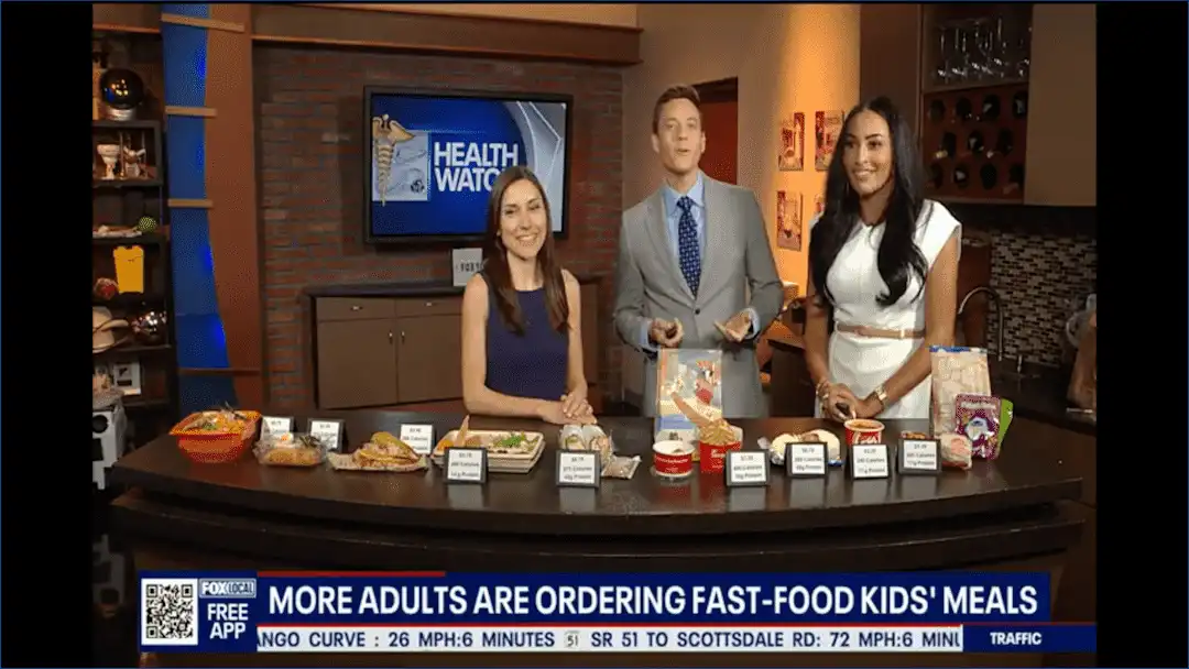 Three TV hosts at Village Clubs Camelback discuss nutrition, showcasing fast-food kids meals under a Health Watch screen.