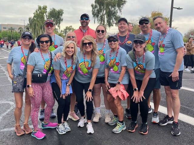 Fourteen adults in blue Village Cares shirts and medals at a Village Clubs Camelback community event, smiling outdoors together.