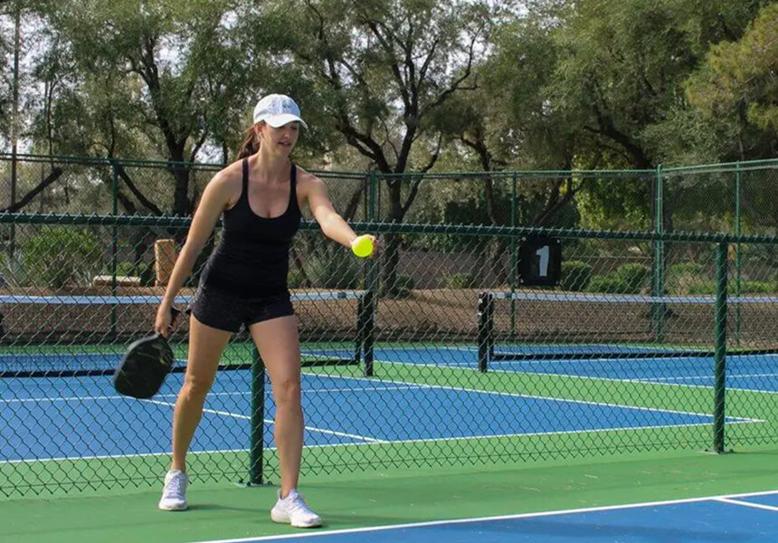 A woman in athletic wear plays pickleball at Village Clubs Camelback’s outdoor court, with trees and a chain-link fence nearby.