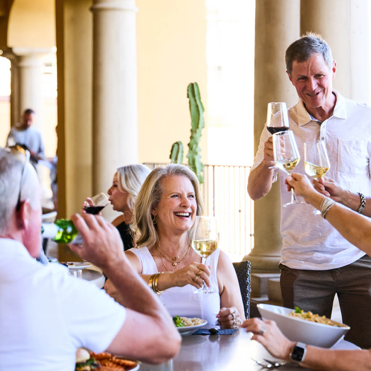 Adults toast and dine outside at Village Clubs Camelback, enjoying Membership Benefits in a sunny setting with columns and cacti.