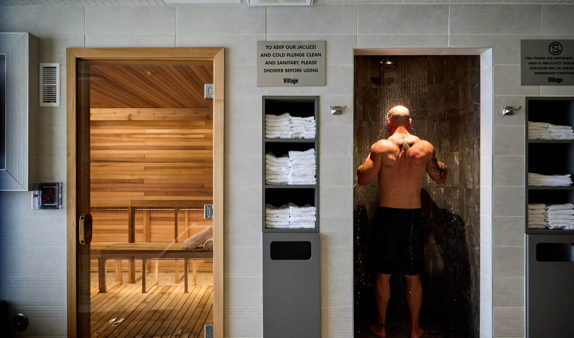 At Village Clubs Camelback, a man showers beside a wooden sauna, with clean towels and reminders for jacuzzi and sauna hygiene.