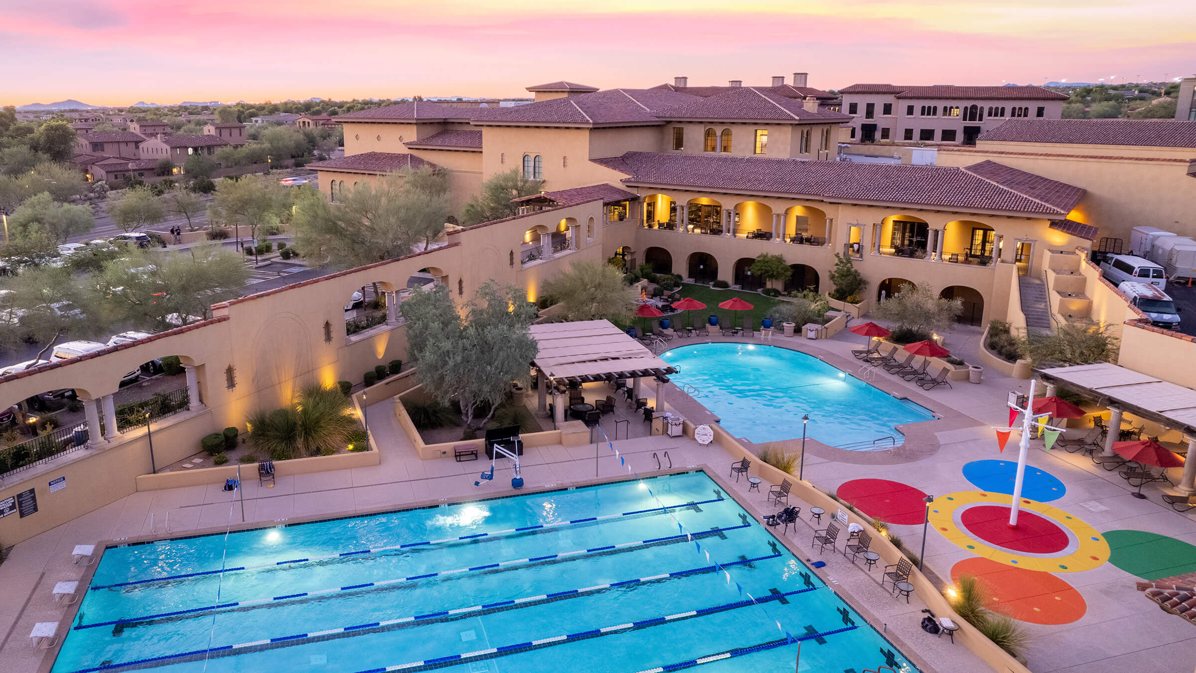 Aerial view of Village Clubs at Camelback with large pools, splash pad, lounge chairs, and Mediterranean-style buildings at sunset.