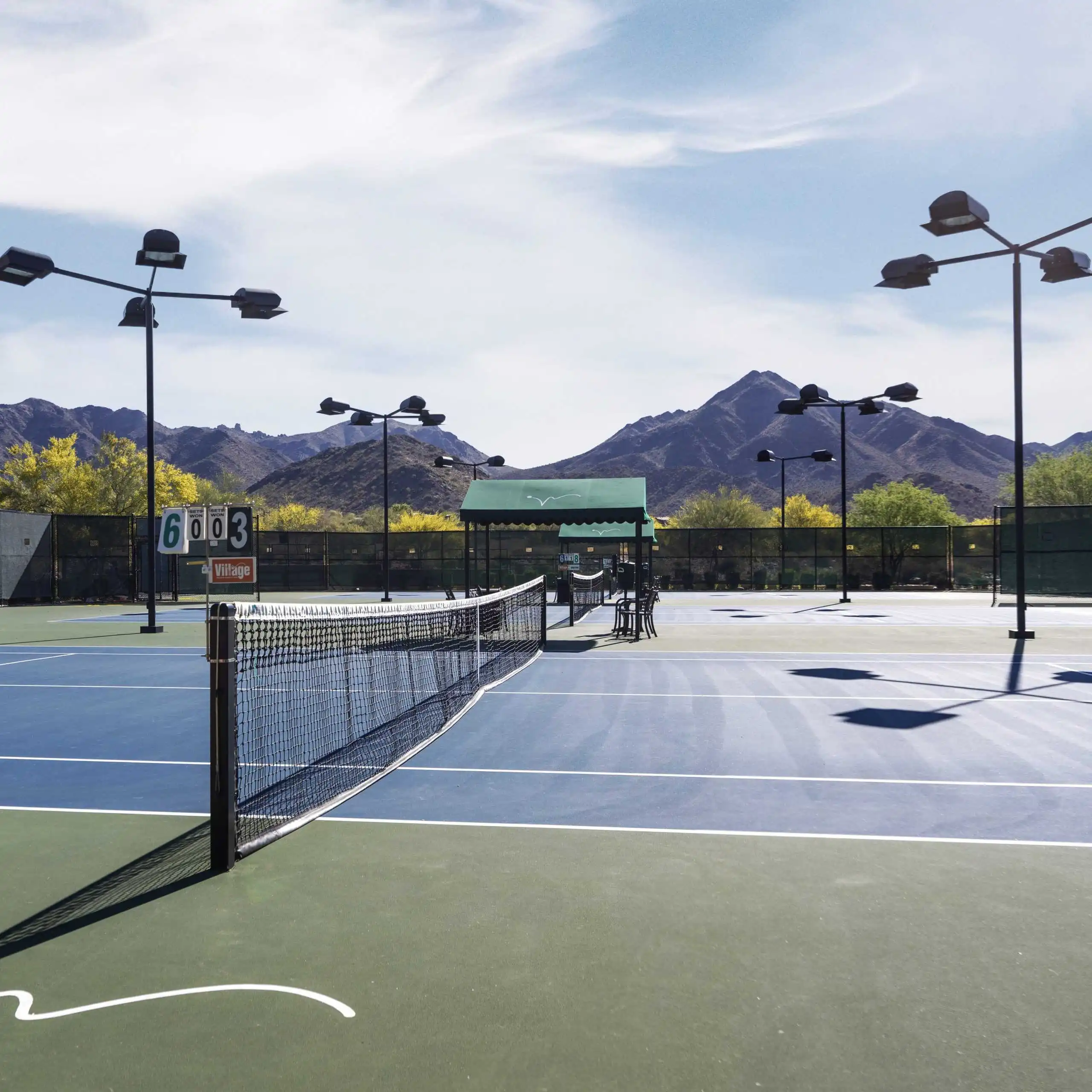 A blue and green tennis court at Village Clubs DC Ranch, with mountains, trees, scoreboards, and eco-friendly lighting for matches.