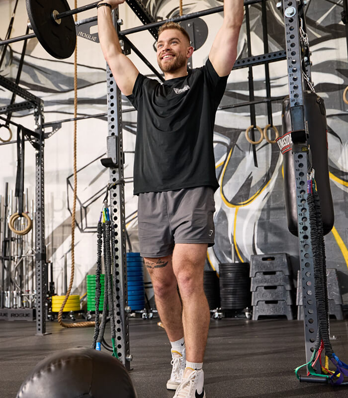 A man lifts a barbell overhead, smiling at Village Clubs Camelback gym in Arizona, with equipment and weights in the background.