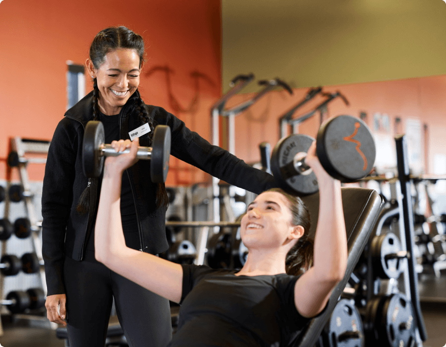 At Village Clubs Camelback, a woman lifts dumbbells on an incline bench with a trainer assisting in a well-equipped gym.