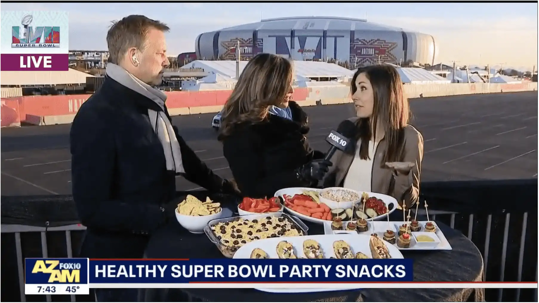 Village Clubs at DC Ranch staff present healthy Super Bowl snacks on TV outdoors, with stadium and event signage in the background.