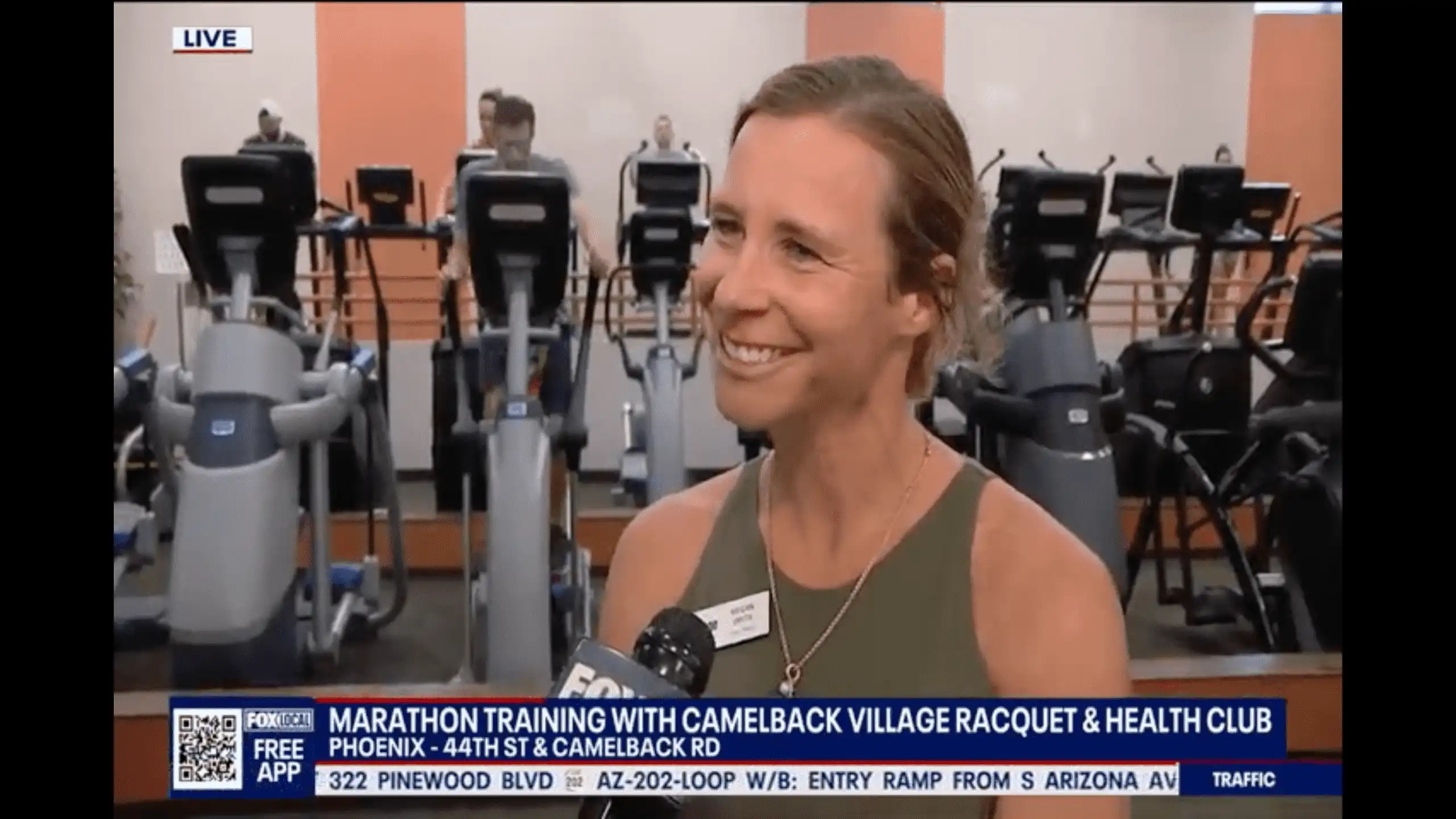 A woman is interviewed at Village Clubs’ Camelback gym, with exercise machines and a news banner on marathon training in view.