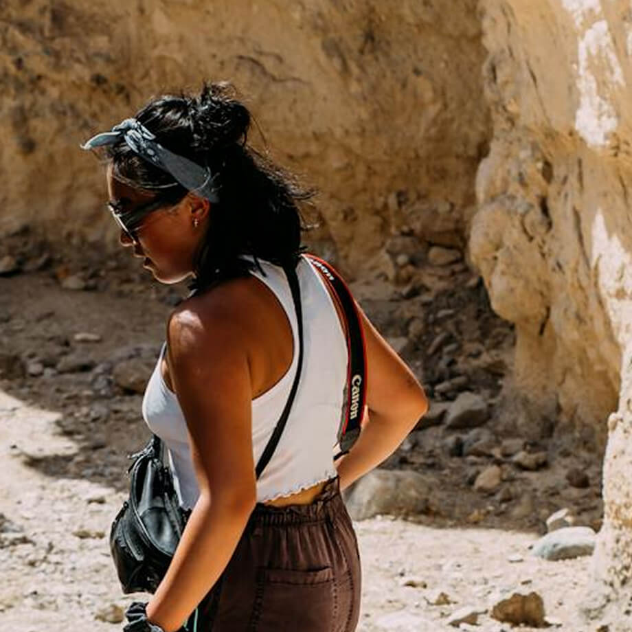 A woman in sunglasses and a white tank top explores a sunlit rocky trail at Village Clubs Camelback, camera strap over her shoulder.