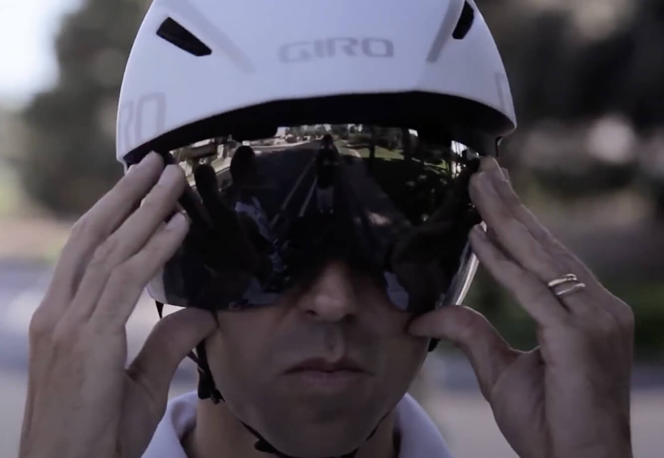 At Village Clubs DC Ranch, a cyclist in a white helmet adjusts reflective sunglasses, mirroring riders and trees in the sun.