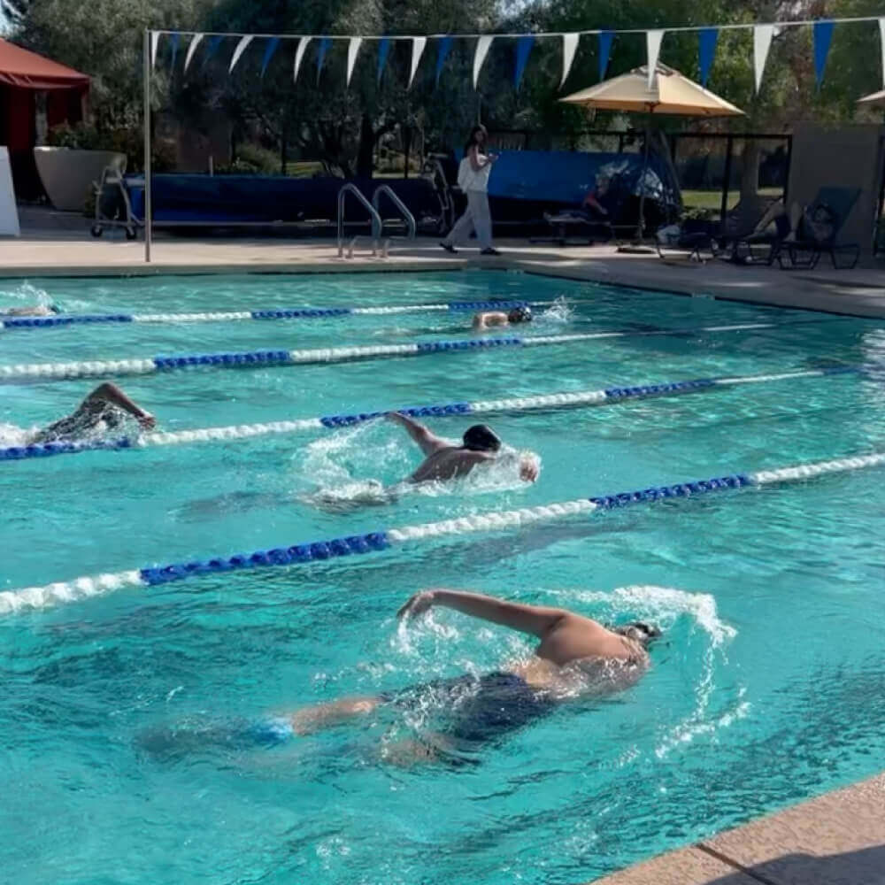 Swimmers practicing freestyle in Village Clubs’ Camelback outdoor pool, with lane dividers, flags, loungers, and umbrellas visible.