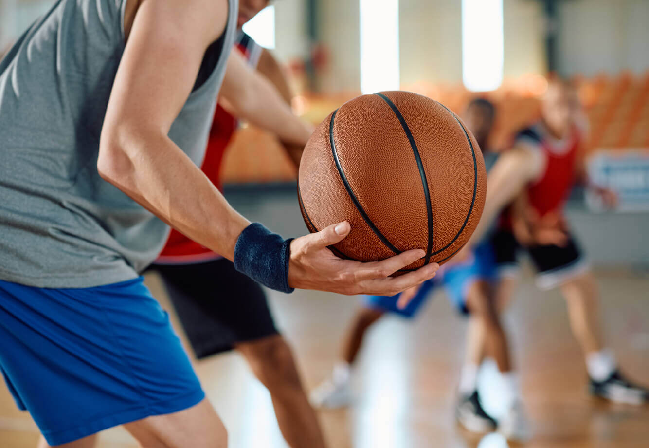 Basketball player at Village Clubs Camelback preparing to pass or dribble on indoor court; others in red, blue jerseys blurred.